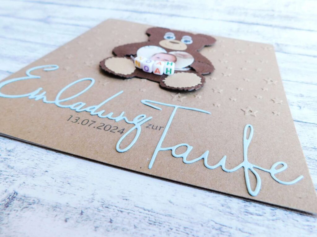 Taufeinladung aus Kraftkarton mit individueller Sternen-Prägung, süßem Teddy-Bären mit Kulleraugen & Foto eines Babys in Herzform darin. Kleine bunte Buchstabenwürfel mit dem Namen des Babys zieren den Teddy. Darunter ist der Schriftzug "Einladung zur Taufe" aus farbigen blauen Papier gestanzt und auf der Taufkarte befestigt. © KARTEN-DESIGN.at | Nicki Heß