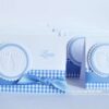 Handgefertigte Tischkarte in weiß mit karierten blauen Karo-Papierstreifen im unteren Bereich mit Taftband darauf. Gepraegte Babyfuesse auf einem gewellten Kreis sind darauf angeordnet. Rechts ist mittig der Gaestename aufgedruckt. Design: First-Lovely-Times ©KARTEN-DESIGN.at