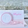 Handgefertigte Tischkarte in weiß mit karierten rosa Karo-Papierstreifen im unteren Bereich mit Taftband darauf. Gepraegte Babyfuesse auf einem gewellten Kreis sind darauf angeordnet. Rechts ist mittig der Gaestename aufgedruckt. Design: First-Lovely-Times ©KARTEN-DESIGN.at